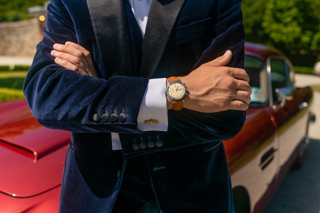 A model poses with an Aston Martin DB5 and his Huguenot Horology vintage inspired gold watch by Watch Brands man in black suit jacket wearing gold round analog watch